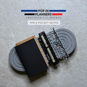 May include: A black notebook with a brown cover, a black pen holder with a white pen, and a clear iridescent ruler. The text reads POP IN PLANNERS PRECISION FIT INSERTS PEN & POCKET NOTES. These items are arranged on a gray oval tray with concentric circles.