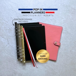 May include: A planner set featuring a coral-colored cover with a snap closure, black inserts with an acrylic-like hard backing, and a metal ring mechanism. Text reads POP IN PLANNERS PRECISION FIT INSERTS, RIBBON BOOKMARK INCLUDED, and ACRYLIC-LIKE HARD BACKING.