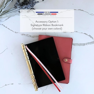 May include: A black planner with a gold-tone metal ring binder mechanism and two thin ribbon bookmarks in red and white. Behind it sits a coral-colored planner cover with a snap closure. The text reads POP IN PLANNERS PRECISION FIT INSERTS.