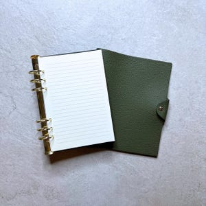 May include: An open, olive green leather binder with gold-colored rings and lined white paper. The binder is partially open, revealing the interior pages. The cover has a button closure. The background is a light gray, textured surface.