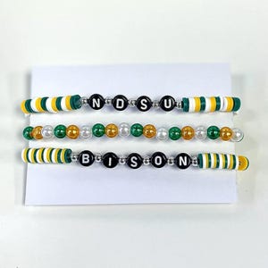 Op de afbeelding: Drie kralenarmbanden met afwisselende groene, witte en gouden kralen. De armbanden spellen "NDSU" en "BISON" in zwarte letters.