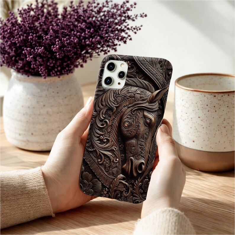 Peut inclure: Une coque de smartphone marron fonc&eacute; pr&eacute;sentant un relief d&eacute;taill&eacute; repr&eacute;sentant une t&ecirc;te de cheval entour&eacute;e de motifs floraux et de volutes complexes. La coque comporte une d&eacute;coupe pour un syst&egrave;me de triple cam&eacute;ra en haut &agrave; gauche.
