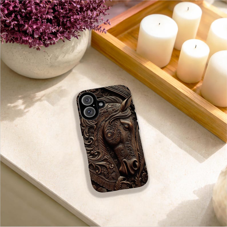 Peut inclure: Une coque de t&eacute;l&eacute;phone marron fonc&eacute; pr&eacute;sentant un relief d&eacute;taill&eacute; repr&eacute;sentant une t&ecirc;te de cheval entour&eacute;e de motifs floraux et de volutes complexes. La coque comporte une d&eacute;coupe pour l'objectif de l'appareil photo et suit la forme rectangulaire.