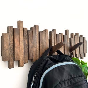May include: A wooden wall-mounted coat rack with a unique design. The rack is made of vertical wooden slats in varying lengths, stained in a dark brown color. A black backpack hangs from the hooks. Green leaves are visible in the corner.