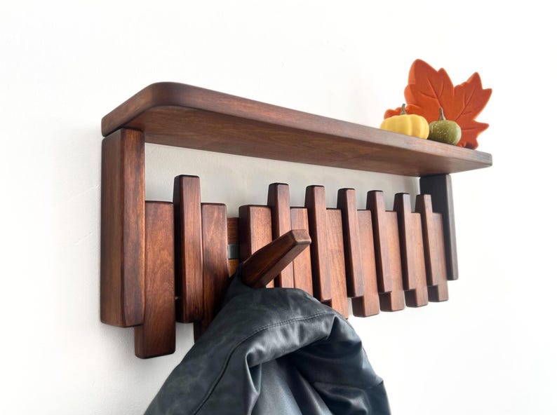 A brown walnut colored piano hook coat rack with a shelf and a black coat hanging from a hook and fall decor on the shelf