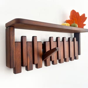 May include: A dark brown wooden wall shelf with a series of vertical pegs that can be used as hooks. The shelf has a flat top and is decorated with a decorative orange leaf, a small yellow gourd, and a green pumpkin.