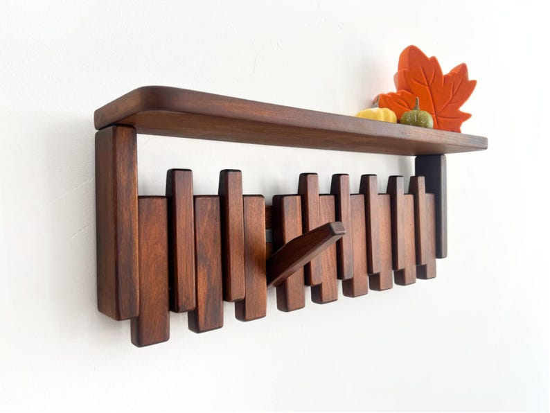 May include: A dark brown wooden wall shelf with a series of vertical pegs that can be used as hooks. The shelf has a flat top and is decorated with a decorative orange leaf, a small yellow gourd, and a green pumpkin.