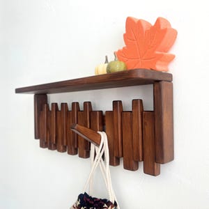 May include: A dark brown wooden wall shelf with a row of hooks below. The shelf is decorated with a large orange leaf and two small gourds. A white bag with a patterned bottom hangs from one of the hooks.