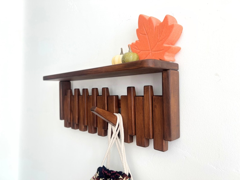 May include: A dark brown wooden wall shelf with a row of hooks below. The shelf is decorated with a large orange leaf and two small gourds. A white bag with a patterned bottom hangs from one of the hooks.