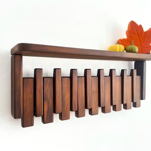 May include: A dark brown wooden wall shelf with a series of vertical wooden slats below. The shelf is decorated with a yellow gourd, a small green gourd, and an orange leaf. The shelf is mounted on a white wall.