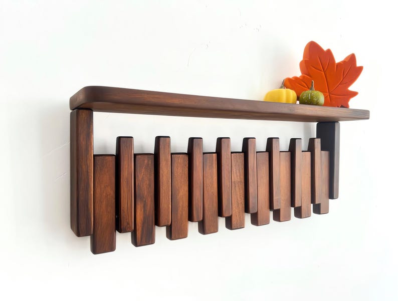 May include: A dark brown wooden wall shelf with a series of vertical wooden slats below. The shelf is decorated with a yellow gourd, a small green gourd, and an orange leaf. The shelf is mounted on a white wall.
