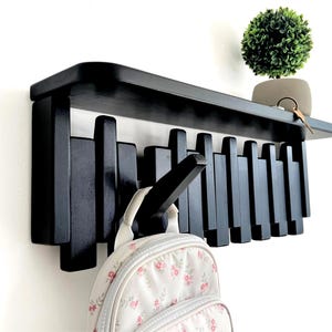 May include: A black wall-mounted coat rack featuring a flat top shelf and a series of vertical wooden slats of varying lengths. One slat extends outward as a hook, currently holding a white backpack with a pink floral pattern.