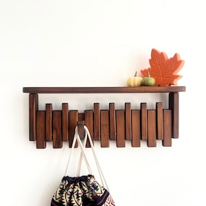 May include: A dark brown wooden wall shelf with vertical peg-like hooks. A patterned drawstring bag hangs from the hooks. The shelf is decorated with a small pumpkin and a fall leaf.