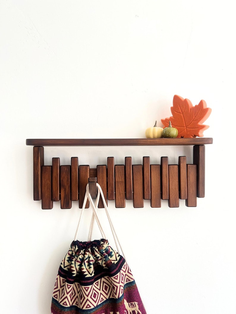 May include: A dark brown wooden wall shelf with vertical peg-like hooks. A patterned drawstring bag hangs from the hooks. The shelf is decorated with a small pumpkin and a fall leaf.