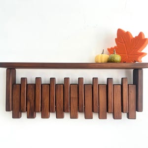 May include: A dark brown wooden shelf with a unique design featuring vertical wooden slats. The shelf is decorated with a large orange leaf and two small pumpkins. The shelf is mounted on a white wall.
