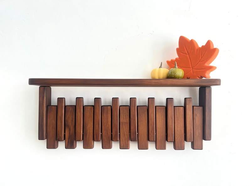 May include: A dark brown wooden shelf with a unique design featuring vertical wooden slats. The shelf is decorated with a large orange leaf and two small pumpkins. The shelf is mounted on a white wall.