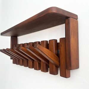 May include: A wooden wall-mounted coat rack with a shelf. The rack is made of dark brown wood and features multiple folding hooks. The shelf is rectangular with rounded corners. The hooks are designed to fold flat against the wall when not in use.