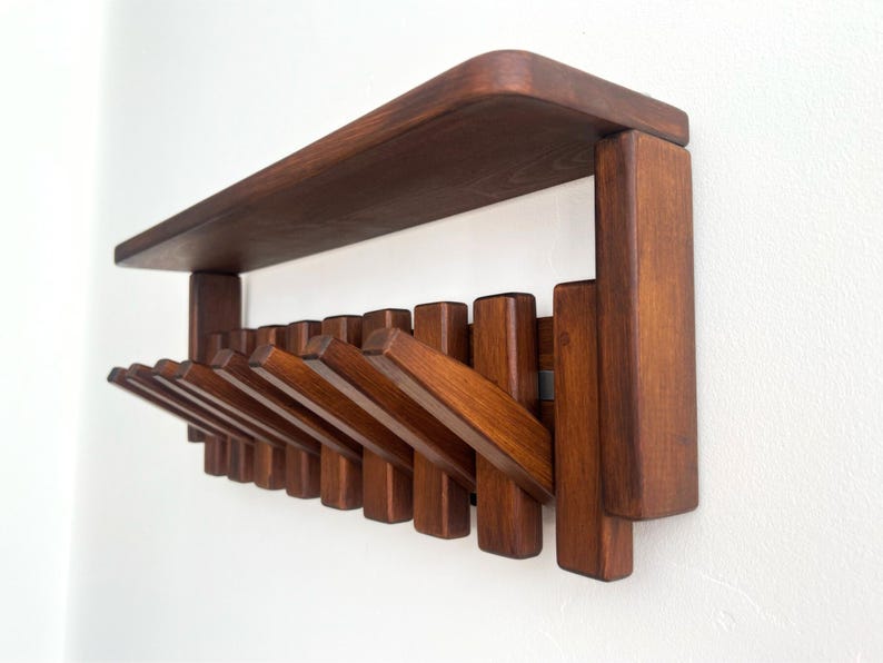 May include: A wooden wall-mounted coat rack with a shelf. The rack is made of dark brown wood and features multiple folding hooks. The shelf is rectangular with rounded corners. The hooks are designed to fold flat against the wall when not in use.