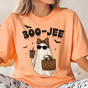 May include: Peach-colored t-shirt with the text "BOO-JEE" and a cartoon ghost wearing sunglasses, a leopard print bow, and carrying a leopard print bag. The ghost is holding a coffee cup. Black bats are scattered around the design. The person is wearing white skull print pants.