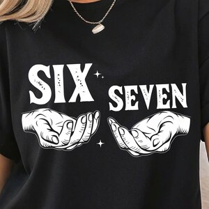 Peut inclure: T-shirt noir avec l'inscription blanche « SIX SEVEN » au-dessus d'un dessin de deux mains ouvertes. Le motif comprend de petits accents d'étoiles. Le t-shirt est de style décontracté.