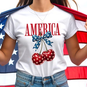 Peut inclure: Un t-shirt blanc à manches courtes avec le texte rouge AMERICA au-dessus d'un motif de deux cerises rouges à étoiles blanches. Les cerises sont reliées par un nœud bleu à motifs d'étoiles blanches. Le t-shirt est porté avec un short en jean bleu.