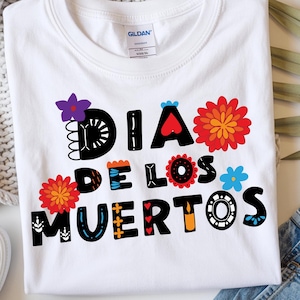 Peut inclure: T-shirt blanc avec l'inscription "DIA DE LOS MUERTOS" en lettres décoratives colorées. Le motif comprend des motifs floraux et en forme de cœur. Le t-shirt est présenté avec un jean, des chaussures et une couverture tricotée.