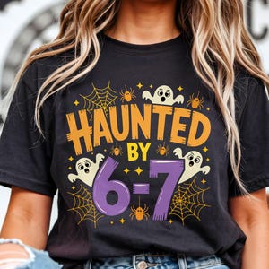 May include: Black t-shirt with a Halloween graphic. The design includes the text "HAUNTED BY 6-7" in orange and purple, with spiderwebs, spiders, and ghost illustrations. The shirt is worn by a person in blue jeans.
