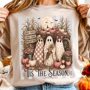 Peut inclure: Un sweat-shirt beige à col rond avec une impression graphique représentant trois fantômes devant une charrette en bois, des citrouilles et des tiges de maïs. Le texte indique GHOST PATCH, CIDER STAND, MOONLIGHT MAZE et Tis The Season.