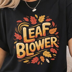 Peut inclure: T-shirt noir avec l'inscription "LEAF BLOWER" dans un design sur le thème de l'automne. Le texte est orange et jaune, entouré de feuilles orange, jaunes et rouges. Le t-shirt a des manches courtes.