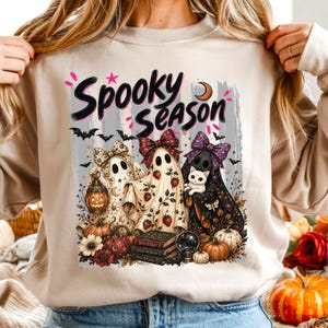 Peut inclure: Un sweat-shirt beige à col rond avec l'inscription "Spooky Season" en lettres noires. Le motif représente trois fantômes portant des nœuds à motifs, tenant une lanterne, un chat blanc, entourés de citrouilles, de fleurs et de livres.