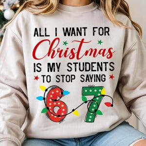 Peut inclure: Un sweat-shirt beige avec le texte "ALL I WANT FOR Christmas IS MY STUDENTS TO STOP SAYING" en noir et rouge. Le chiffre "67" est décoré de guirlandes lumineuses. Un sapin de Noël est en arrière-plan.