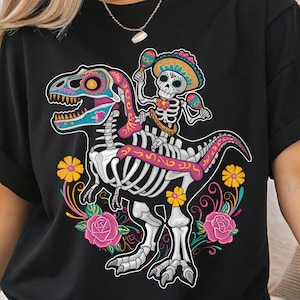 Peut inclure: T-shirt noir avec un motif du Jour des Morts. Un squelette portant un sombrero chevauche un dinosaure orné de motifs colorés. Le motif comprend des maracas, des fleurs et des roses.