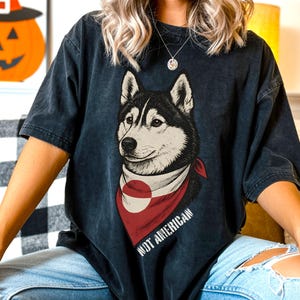 Peut inclure: T-shirt gris foncé avec une illustration de chien husky portant un bandana rouge avec un cercle blanc. Le texte "NOT AMERICAN" est imprimé sur le bandana. Une décoration de citrouille d'Halloween est visible en arrière-plan.