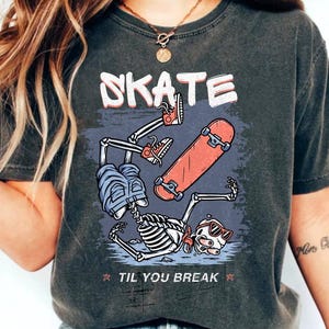 Peut inclure: T-shirt gris foncé avec un motif de squelette faisant du skateboard. Le mot "SKATE" est au-dessus du motif et "TIL YOU BREAK" en dessous. Le skateboard est rouge et le sac à dos est bleu.