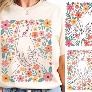 Peut inclure: T-shirt crème avec un motif floral. Le motif représente une main de mère tenant les mains de deux enfants, avec le mot "Mama" sur la main de la mère. Les fleurs sont roses, oranges et jaunes.