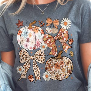 Peut inclure: T-shirt gris avec un motif sur le thème de l'automne. Le design comprend des citrouilles, des nœuds, des marguerites et des feuilles d'automne. Les citrouilles et les nœuds ont des motifs floraux et léopard. Style de t-shirt décontracté.