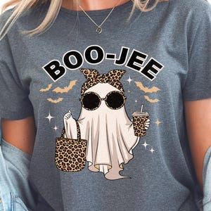 Peut inclure: Un t-shirt gris avec un motif sur le thème d'Halloween. Le motif représente un fantôme portant des lunettes de soleil et un nœud léopard, tenant un sac et une boisson. Le texte "BOO-JEE" est au-dessus du fantôme.