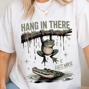 Puede incluir: Camiseta blanca con un diseño de dibujos animados. Una rana colgada de una rama con el texto "HANG IN THERE". Debajo, un caimán en el agua y el texto "IT GETS WORSE".