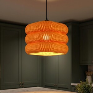 Op de afbeelding: Een feloranje hanglamp met een cilindrische, drielaagse vorm en een fijne, honingraatachtige gaasstructuur. De lamp hangt aan een dun zwart snoer en verspreidt een warme, zachte gloed vanuit het midden die de ruimte eronder verlicht.