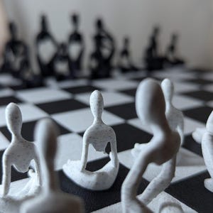 May include: A unique chess set featuring white, abstract human-like figures on a black and white checkered board. The pieces are in various poses, with some seated and others standing. The background shows the blurred black chess pieces.
