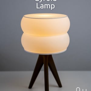 Op de afbeelding: Een moderne tafellamp met de tekst "Gyroid Lamp". De lamp heeft een gestapeld, afgerond ontwerp met een witte kap en een donkerbruine driepootbasis. De lamp is verlicht en straalt een warme gloed uit.