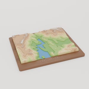 May include: A 3D topographic map of Lake Waramaug, Connecticut, featuring layered elevation contours in shades of blue, green, and tan. The map is mounted on a rectangular wooden base with the text Lake Waramaug engraved on the front.