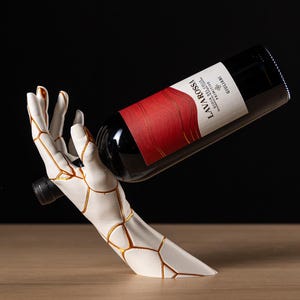 May include: A white ceramic wine bottle holder in the shape of a hand, with gold accents. The hand cradles a red wine bottle with a label that reads "LAVAROSSA". The holder is on a wooden surface against a black background.