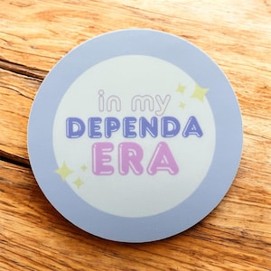 May include: A round sticker with a light blue border and the text "in my DEPENDA ERA" in purple and pink bubble letters. Yellow star accents are scattered around the text. The sticker is on a wooden surface.