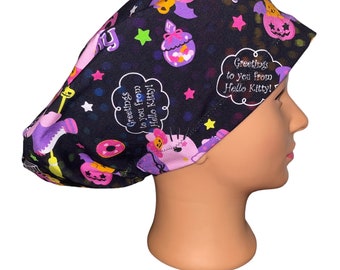May include: A black surgical scrub cap featuring a Hello Kitty Halloween theme. The cap is covered in colorful illustrations of Hello Kitty characters, pumpkins, stars, and the text "Greetings to you from Hello Kitty!"