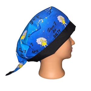 May include: Blue surgical scrub hat with a black elastic band. The hat has a repeating pattern of a cartoon girl holding an umbrella and the text "Don't be salty".