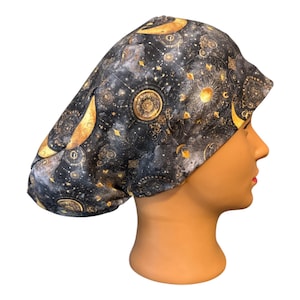May include: A scrub cap featuring a dark gray and black fabric with a celestial pattern of gold moons, suns, and stars. The head covering has a rounded, gathered shape designed to fit securely over the head with a patterned print throughout.
