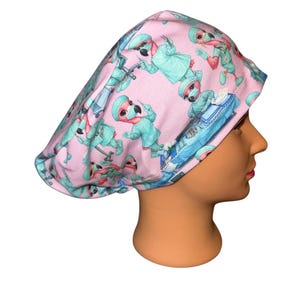May include: A pink surgical scrub cap featuring a repeating pattern of cartoon flamingos dressed as surgeons. The flamingos are wearing surgical masks and scrubs in shades of teal and blue. The cap is designed to cover the hair.