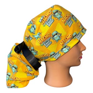 May include: A yellow scrub cap featuring a repeating pattern of cartoon nachos wearing sunglasses and surgical masks, with the text "Nacho Average First Assist" printed in bold letters. The cap includes a black satin ribbon tie at the back.