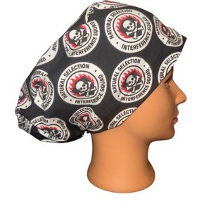 May include: Black surgical scrub cap with a repeating circular pattern. Each circle features a skull and crossbones design with the words "Natural Selection" and "Interference Squad". The cap is worn on a mannequin head.